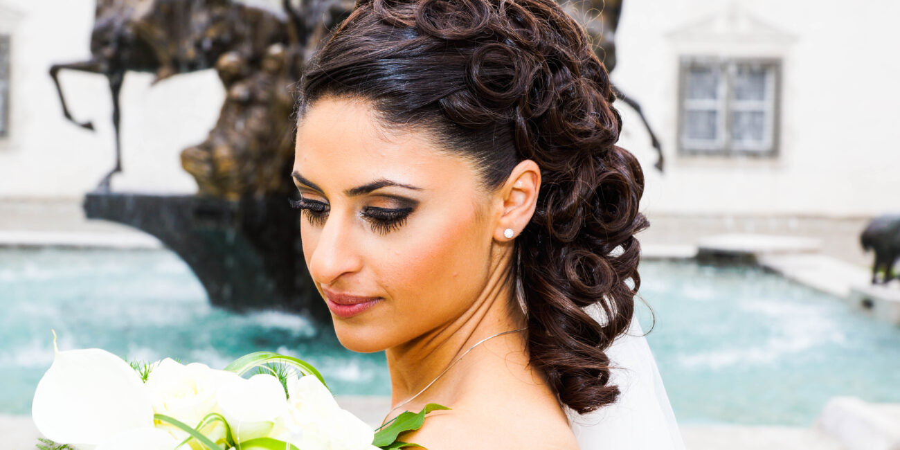 Strahlende Braut mit elegant gestylten Locken und glamourösem Make-up, die ihren Blumenstrauß hält, vor einem kunstvollen Springbrunnen.