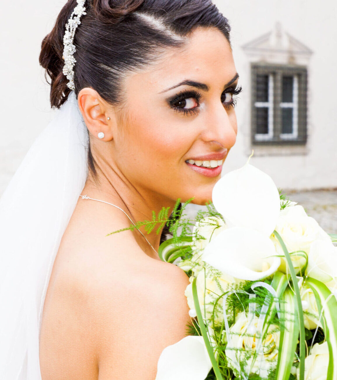 Strahlende Braut mit elegantem Hochsteckfrisur und funkelndem Haarschmuck, hält einen üppigen Brautstrauß aus weißen Calla-Lilien und grünen Akzenten.
