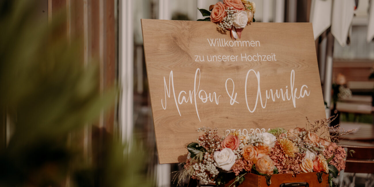 Rustikales Hochzeitsschild aus Holz mit weißer Kalligraphie, umrahmt von Blumen in warmen Tönen. Begrüßung zur Hochzeit von Marlon & Annika.