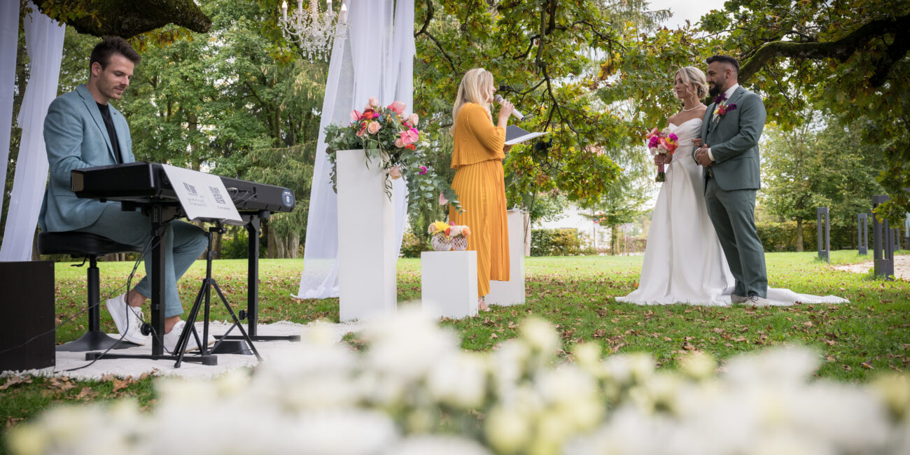 Emotionale freie Trauung unter einem Baum mit Brautpaar, Traurednerin und Live-Musiker am Piano, umgeben von stilvoller Dekoration im Freien.